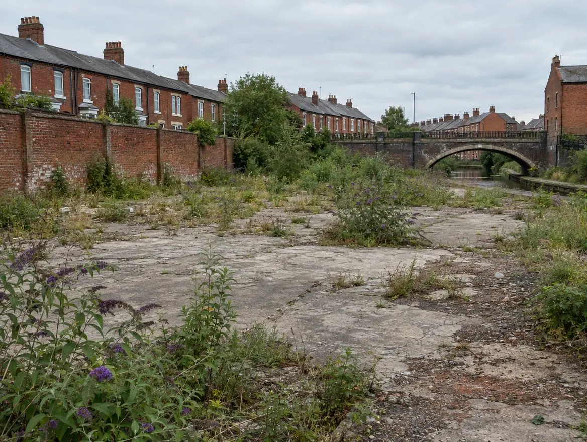 Cleared former industrial site with cracked concrete and weeds beside terraced houses