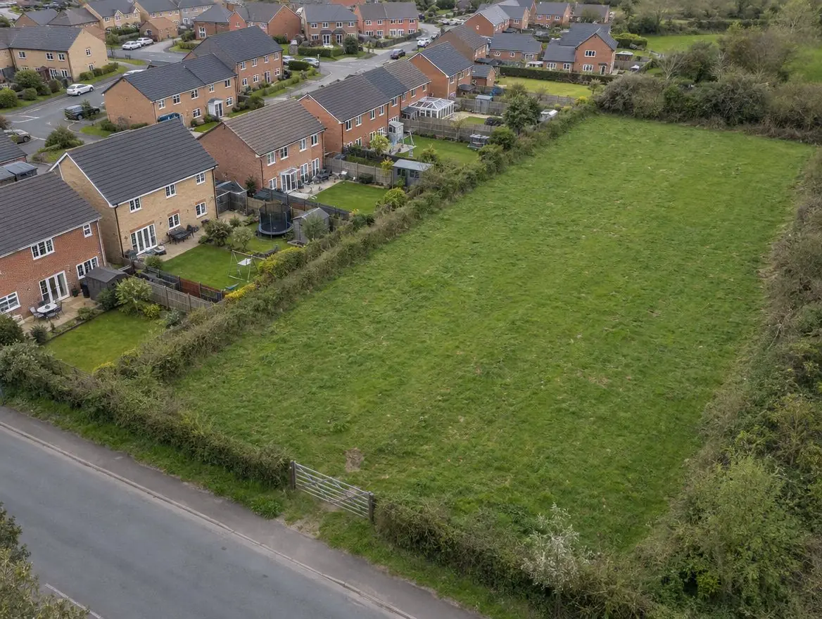Grass field beside the back gardens of a modern housing estate, with a field gate onto the road