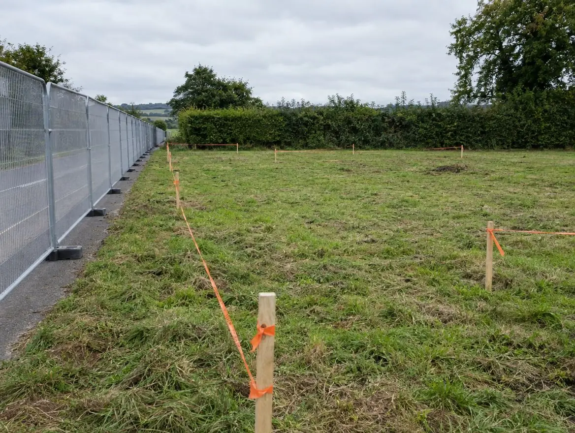 Greenfield site with timber setting-out pegs and orange tape beside a temporary mesh fence